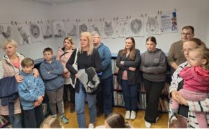 Skupina odraslih in otrok stoji v knjižnici ali učilnici ob knjižnih policah, na steni za njimi pa so razstavljene risbe živali, pripete z lesenimi ščipalkami. Ljudje so oblečeni v vsakdanja oblačila, večina gleda v isto smer, kot da poslušajo predstavitev; ena oseba drži majhnega otroka v naročju. Atmosfera je mirna in pozorna.