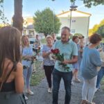 Skupina obiskovalcev pred Knjižnico Ivančna Gorica; nekateri v rokah držijo rdeče vrtnice in kozarce ter se družijo ob dogodku na prostem.
